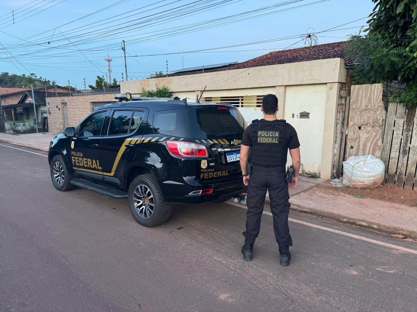 A imagem em destaque mostra um policial federal próximo a uma viatura da instituição e a um dos locais alvos dos mandados.